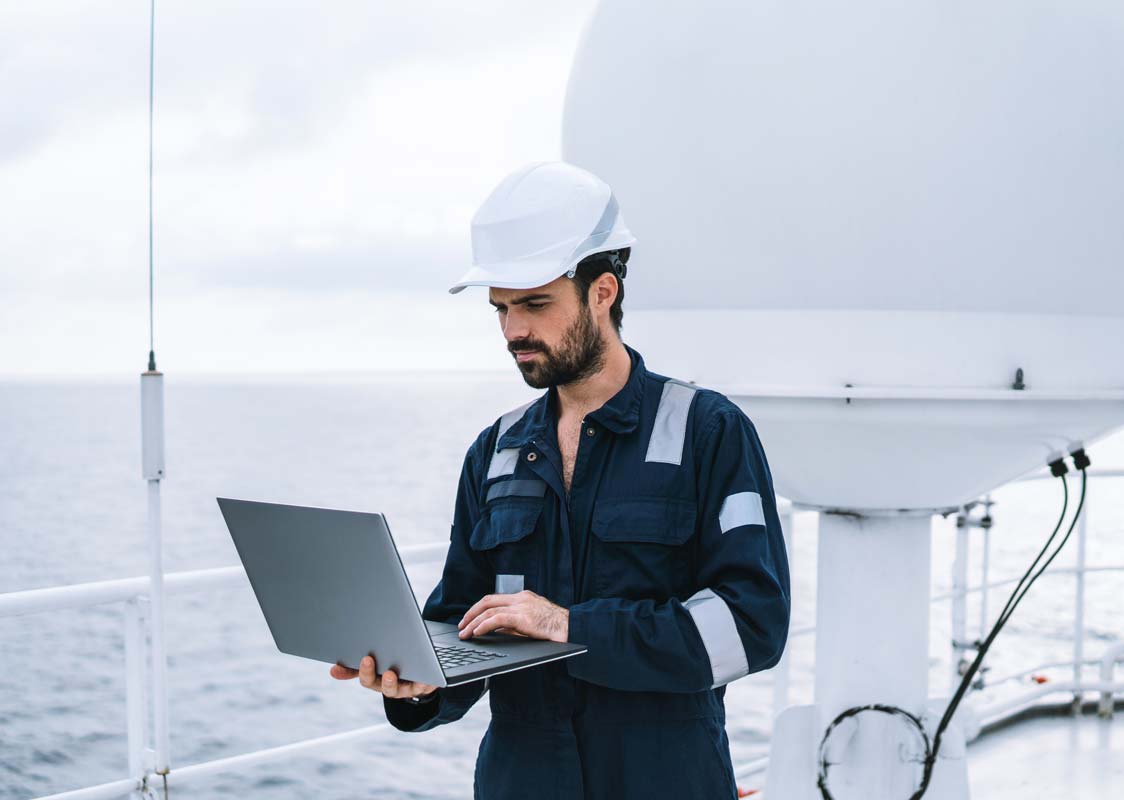 Marine service technician or serviceman near VSAT terminal on deck of vessel or ship. He is working on laptop or notebook